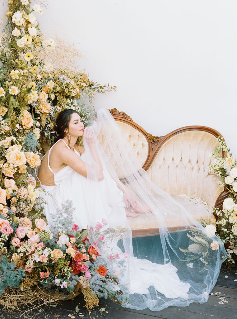 A large floral arrangement made of yellow, blue, and pink wildflower blooms, surrounding an antique, yellow velvet sofa. A model wearing a flow-y, white wedding dress sits on the sofa. She is holding her knees to her chest, looking off into the distance, and smiling. Photographed by Kim Branagan at Nectar + Bloom in San Diego, CA.