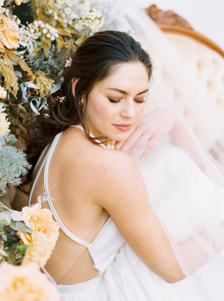 Close up side-view shot of a woman wearing a white wedding dress sitting on a vintage couch, with a wall of yellow, white, and blue flowers to her left.