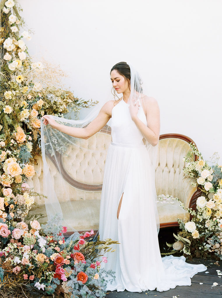 A large floral arrangement made of yellow, blue, and pink wildflower blooms, surrounding an antique, yellow velvet sofa. A model wearing a flow-y, white wedding dress and long veil stands in front of the sofa. She is looking down at the ground and smiling. Photographed by Kim Branagan at Nectar + Bloom in San Diego, CA.