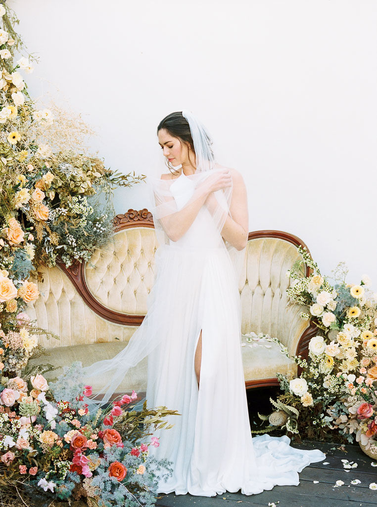A large floral arrangement made of yellow, blue, and pink wildflower blooms, surrounding an antique, yellow velvet sofa. A model wearing a flow-y, white wedding dress and long veil stands in front of the sofa. She is looking down at the ground, wrapping her veil around herself, and smiling. Photographed by Kim Branagan at Nectar + Bloom in San Diego, CA.