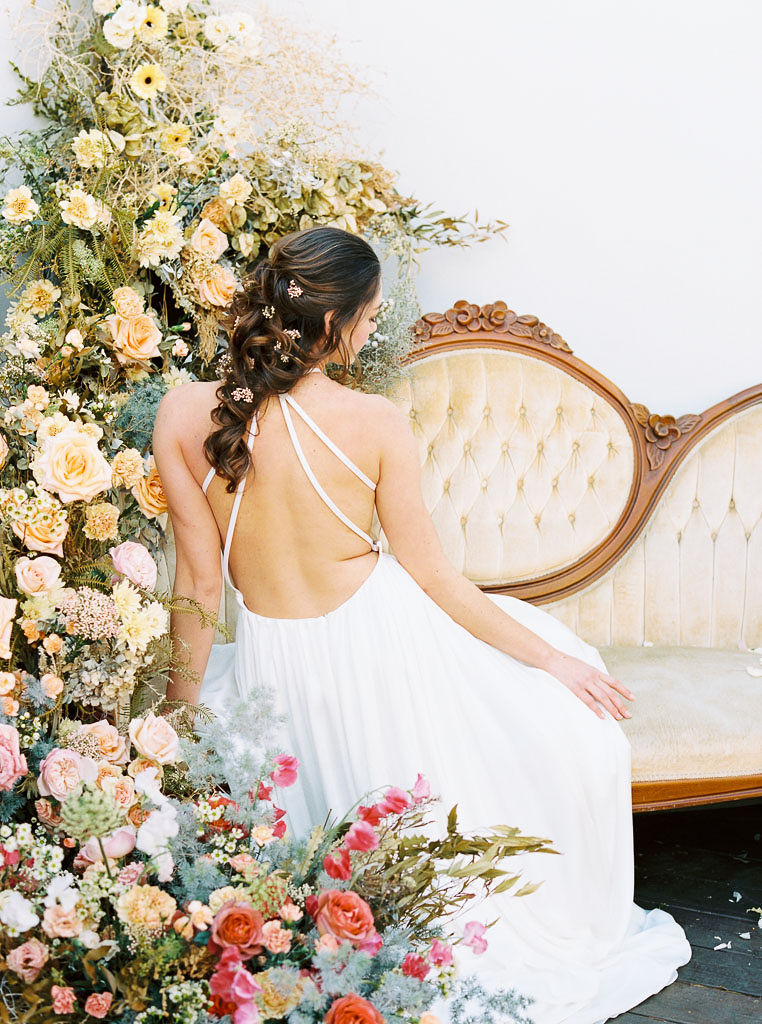 A large floral arrangement made of yellow, blue, and pink wildflower blooms, surrounding an antique, yellow velvet sofa. A model wearing a flow-y, white wedding dress sits on the sofa. Her back is facing the camera. Her long brown hair cascades down her back. Photographed by Kim Branagan at Nectar + Bloom in San Diego, CA.