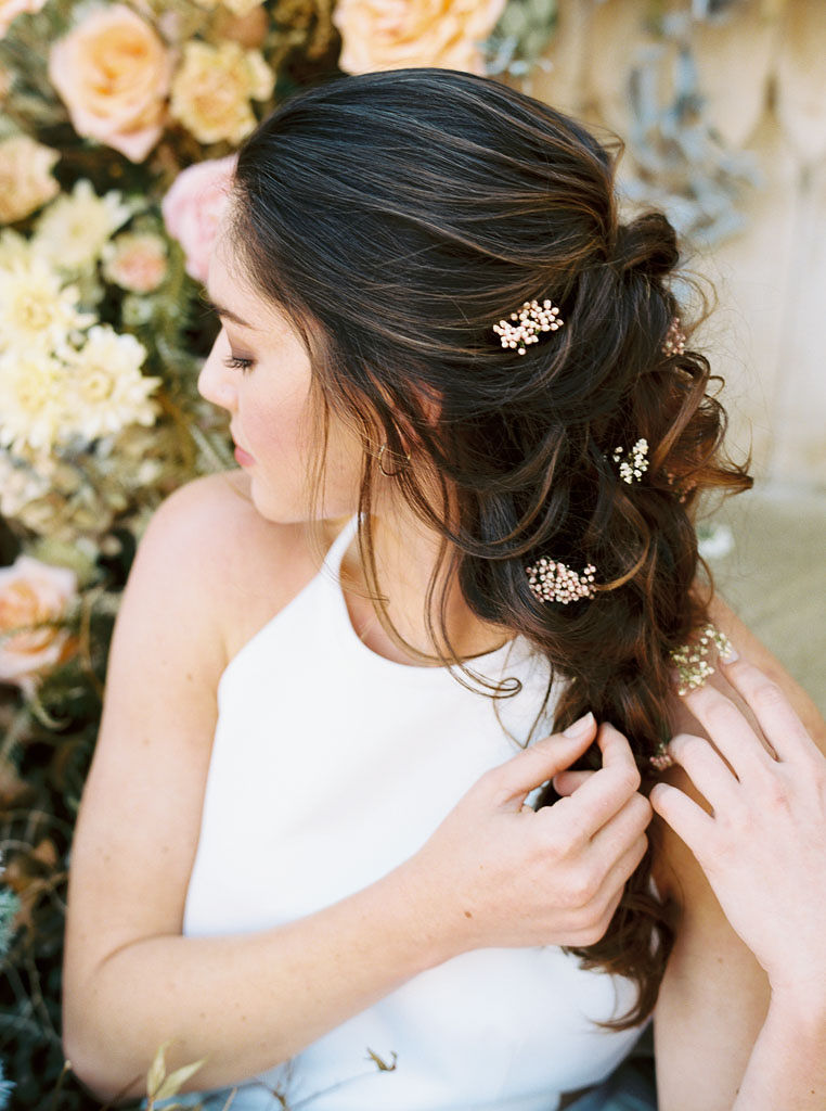 Close up shot of a model wearing a white wedding dress with her face turned away from the camera. Her long brown hair is done in a loose braid, with sprigs of white wildflowers woven into the braid. Photographed by San Diego wedding photographer Kim Branagan.