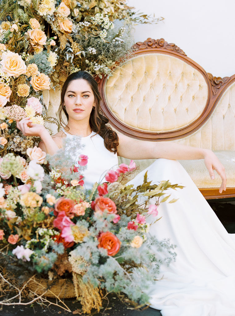 A large floral arrangement made of yellow, blue, and pink wildflower blooms, surrounding an antique, yellow velvet sofa. A model wearing a flow-y, white wedding dress and long veil sits on the ground in front of the sofa. She is looking straight at the camera and holding a flower from the arrangement. Photographed by Kim Branagan at Nectar + Bloom in San Diego, CA.