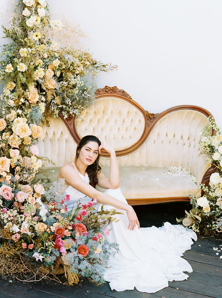 A large floral arrangement made of yellow, blue, and pink wildflower blooms, surrounding an antique, yellow velvet sofa. A model wearing a flow-y, white wedding dress and long veil sits on the ground in front of the sofa. She is looking straight at the camera, with her elbow resting on her knee and her hand resting on her forehead. Photographed by Kim Branagan at Nectar + Bloom in San Diego, CA.