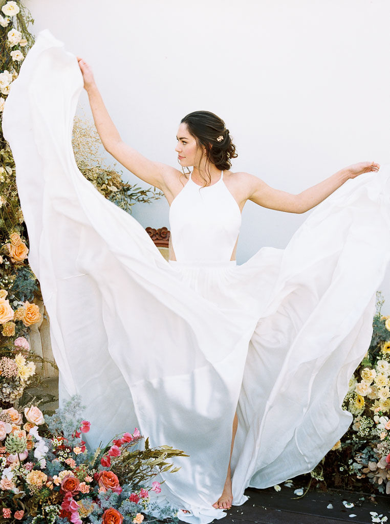 A model wearing a flowing white wedding dress with the skirt of the dress blowing in the air around her. A floral installation of colorful wildflowers surrounds her. Photographed for a floral workshop at Nectar + Bloom by San Diego editorial and wedding photographer Kim Branagan.