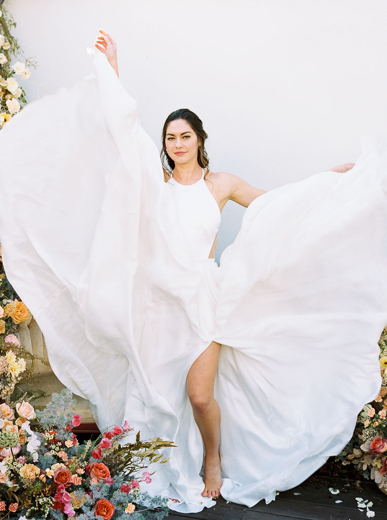 A model wearing a flowing white wedding dress with the skirt of the dress blowing in the air around her. A floral installation of colorful wildflowers surrounds her. Photographed for a floral workshop at Nectar + Bloom by San Diego editorial and wedding photographer Kim Branagan.