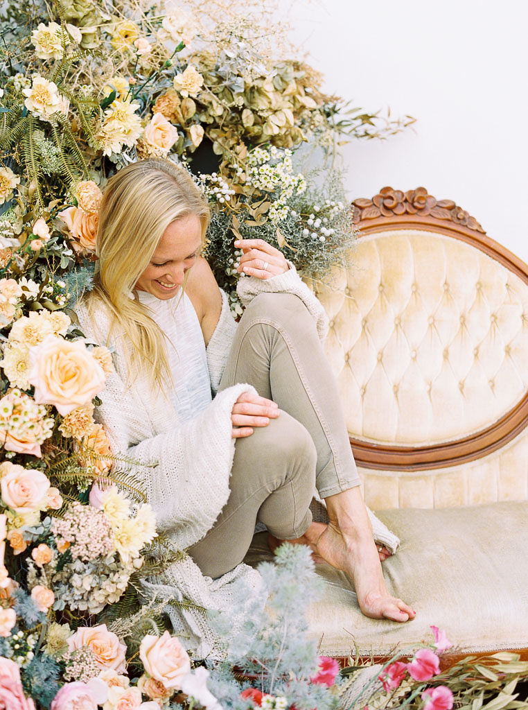 Jen, owner of Nectar + Bloom sits on a yellow, crushed velvet couch and looks down at the ground, laughing. She is sitting next to the vintage, wildflower floral installation she designed. Photographed by San Diego commercial photographer Kim Branagan.