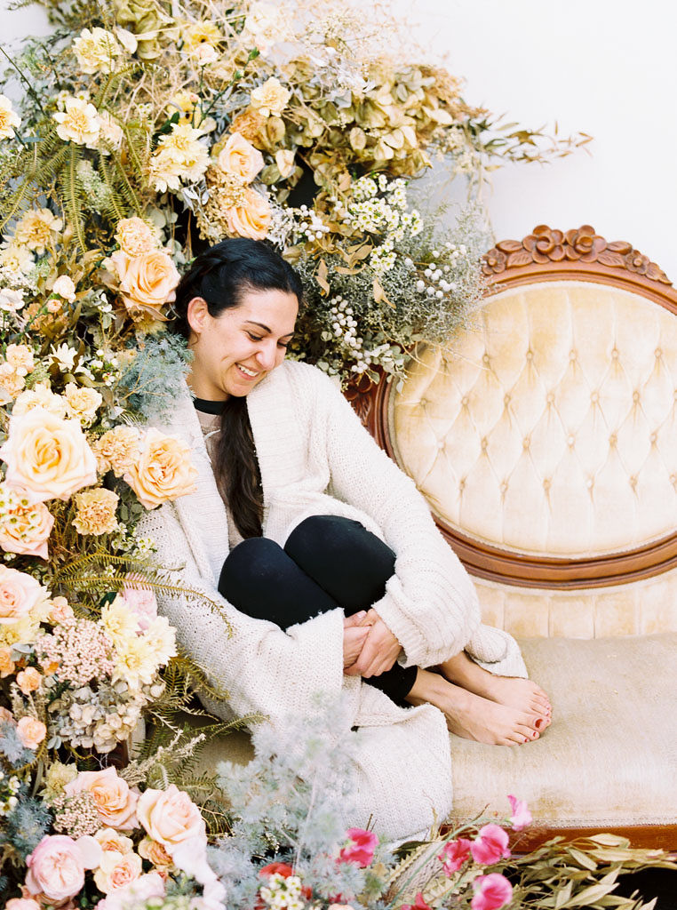 Dena, who attended a floral workshop at Nectar + Bloom, sits on a yellow, crushed velvet couch next to the vintage, wildflower floral installation she designed with Jen, owner of Nectar + Bloom in San Diego, CA.