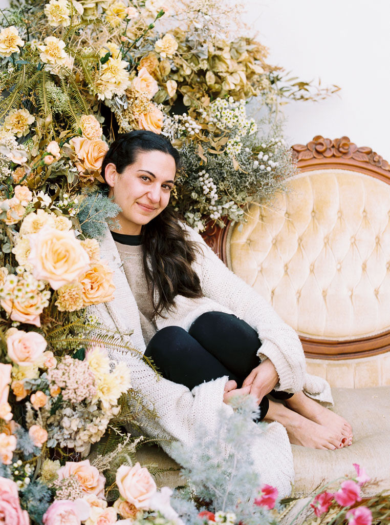 Dena, who attended a floral workshop at Nectar + Bloom, sits on a yellow, crushed velvet couch next to the vintage, wildflower floral installation she designed with Jen, owner of Nectar + Bloom in San Diego, CA. Dena is smiling and looking directly at the camera.