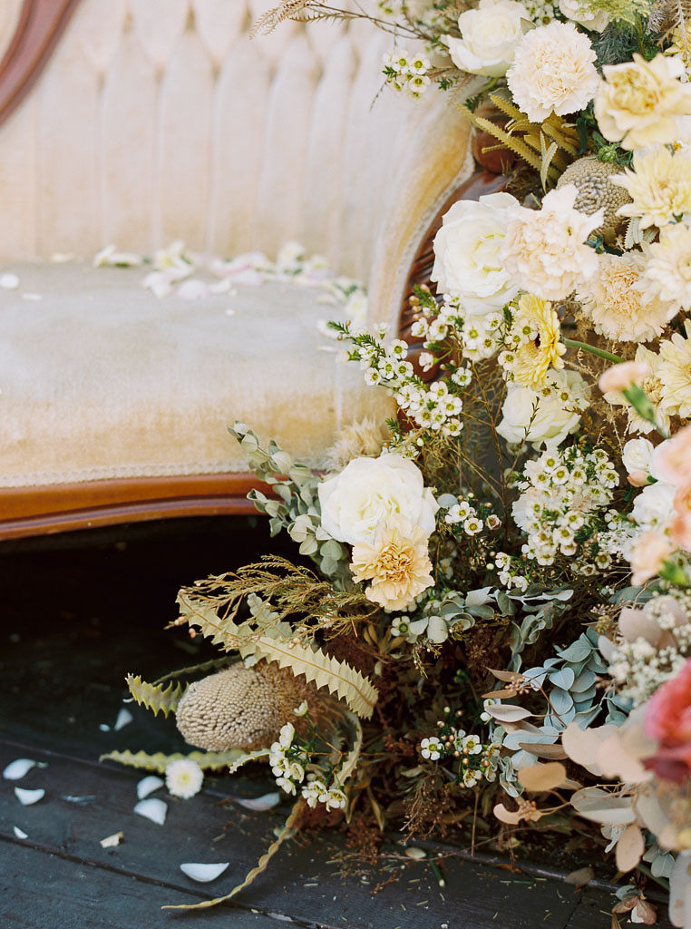 A large floral arrangement of yellow, cream, and pink flowers with lots of greenery next to a gold colored couch.