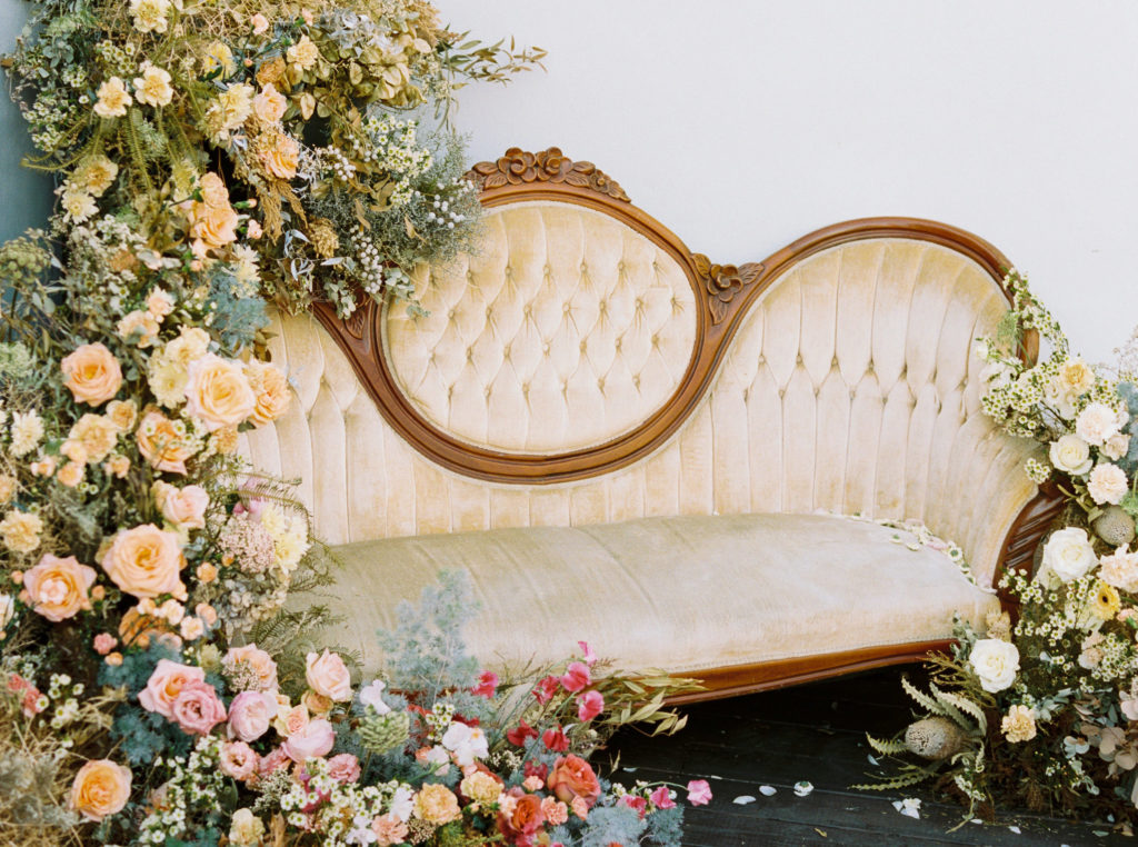 A large floral arrangement made of lush wildflower blooms and vibrant spring colors, surrounding an antique, yellow velvet sofa. Photographed by Kim Branagan at Nectar + Bloom in San Diego, CA.