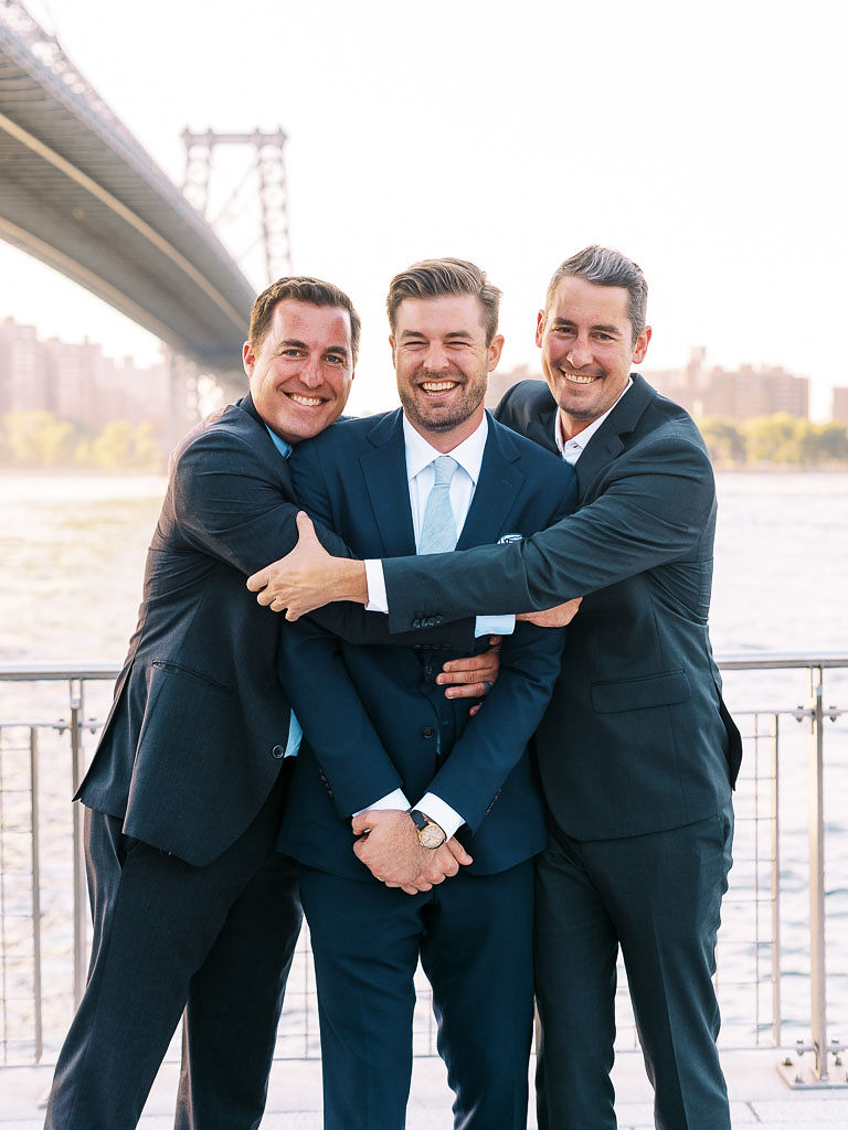 A man and his friends, all wearing suits, embrace and laugh together on the man's wedding day