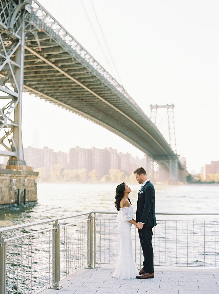 A bride and groom stand facing each other, holding hands, in front of the Williamsburg bridge on their wedding day. Photo by New York City wedding photographer Kim Branagan