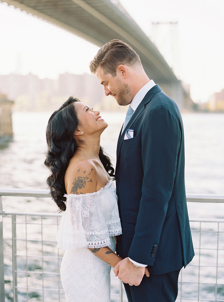 A bride and groom gaze into each other's eye, smiling and holding hands, with the Williamsburg bridge in the in background