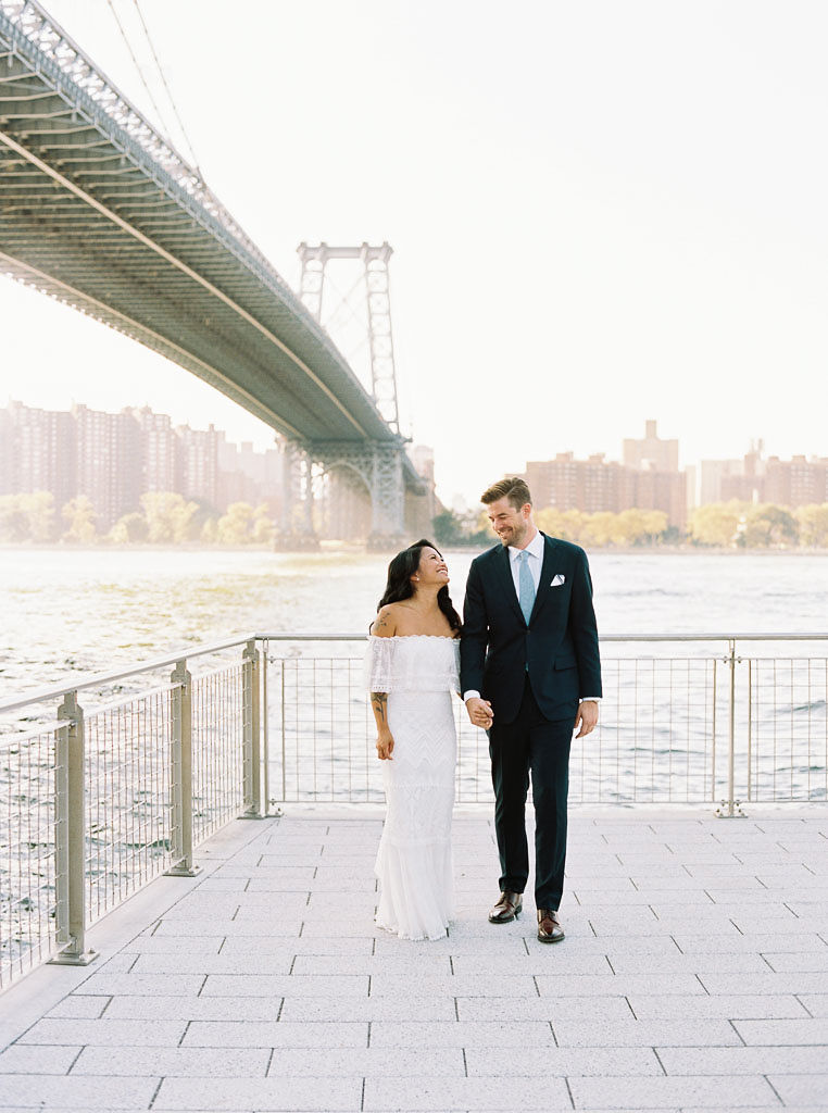 A man and woman stand on a walkway near the water in Brooklyn, NY on their wedding day. They are smiling at each other and walking hand in hand.