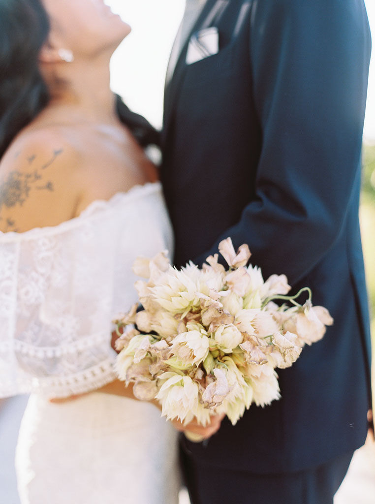 A couple wearing their wedding clothes embrace on their wedding day. The bride is holding a blush and white bouquet of flowers.