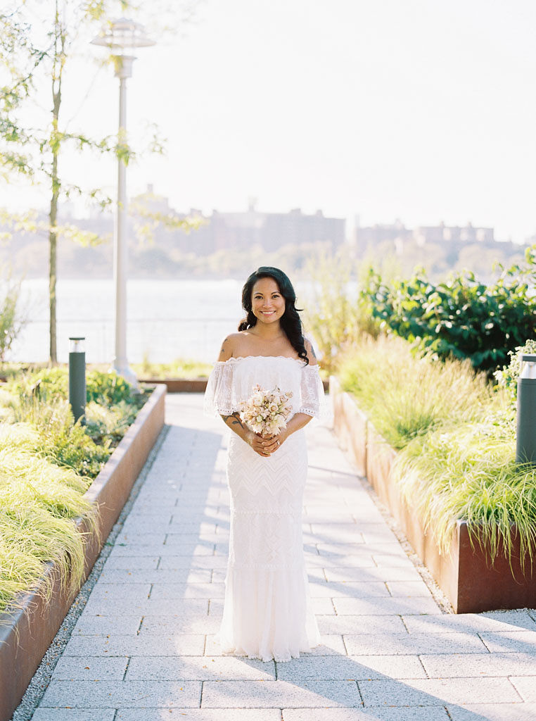 A bride looks directly at the camera, smiling and holding her bridal bouquet. The Manhattan skyline is in the background. Taken by East Coast Wedding Photographer Kim Branagan.