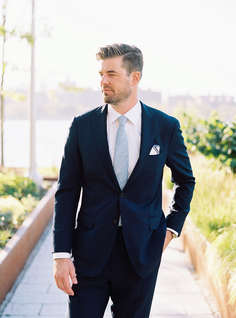 A man wears a suit on his wedding day, his hand in his pocket. He is looking off to the side. The Manhattan skyline is in the background.