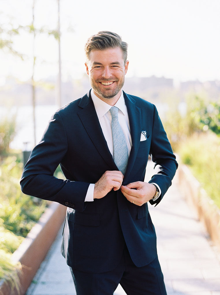 A man smiles at the camera as he buttons his suit coat on his wedding day.