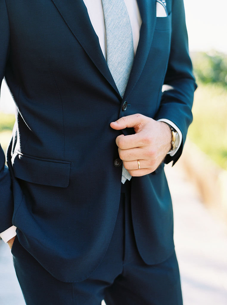 Close up shot of a man buttoning his suit coat on his wedding day. His gold wedding band is visible on his hand.