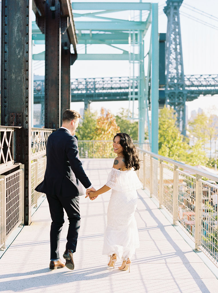 A couple walks in Domino Park on their wedding day. Their backs are to the camera, but the bride is looking back over her shoulder at the camera and laughing. Taken by New York wedding photographer Kim Branagan.