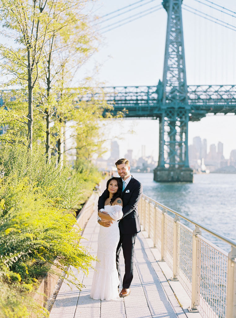 A couple stands in front of the Williamsburg bridge on their wedding day. The bride is standing in front of her husband, leaning against him, and he has his arms around her.