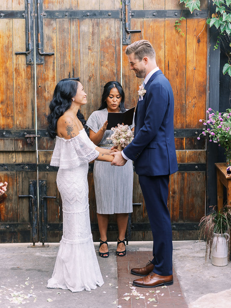 A bride and groom smile at each other and hold hands during their wedding ceremony. Their, modern, urban, boho wedding was held at Aurora restaurant in Brooklyn, NY.