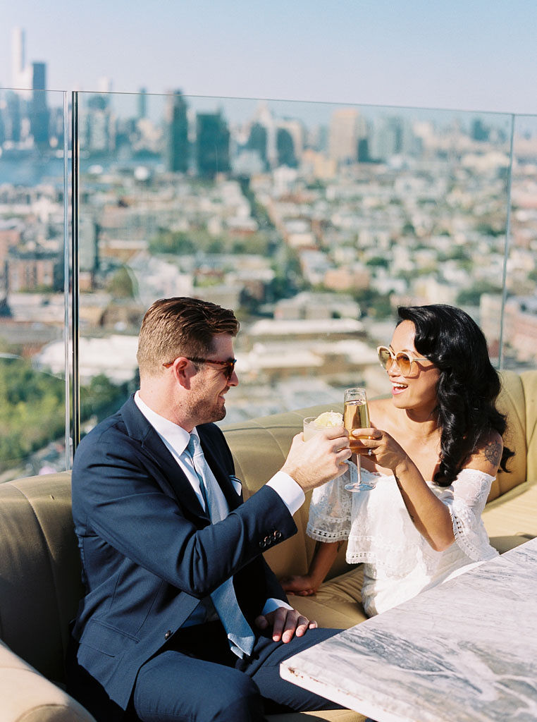 A bride and groom toast each other as they share a cocktail before their wedding ceremony. They are sitting at a rooftop bar in Williamsburg, NY.