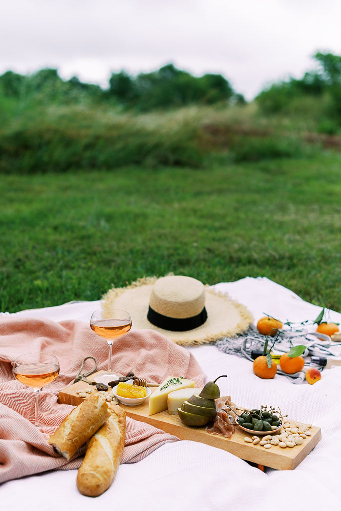 A charcuterie board sits on a pink picnic blanket along with a straw hat. With fruit, olives, cheese, meat, and nuts and a bottle of rose. Photographed by Washington DC brand photographer Kim Branagan.