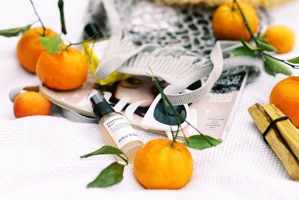A white woven bag, oranges, luxury clean bueaty products, and a magazine laying on a white picnic blanket.