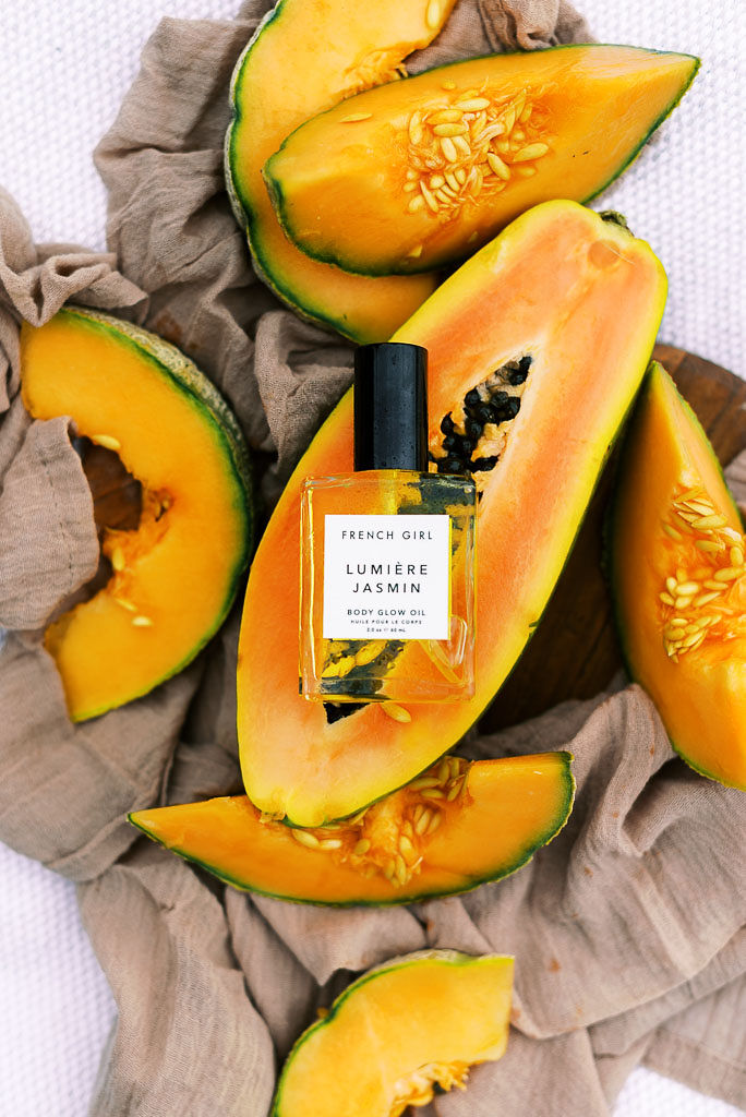 A small bottle of a luxury beauty product from Verde Lusso's summer beauty box laying on top of cut open papayas on a mauve picnic blanket.
