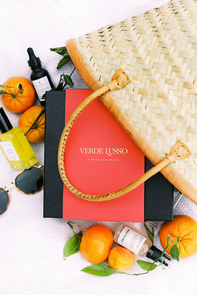 Close up shot of Verde Lusso beauty box with the company logo on the front of the box, laying next to a wicker tote bag, with oranges and the beauty products surrounding the bag. Photographed by Washington DC product photographer Kim Branagan.