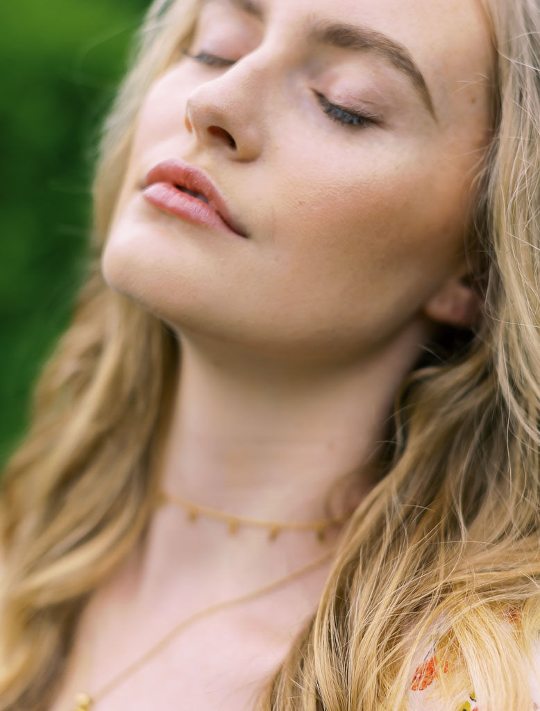 Close up shot of a woman tilting her head back with her eyes closed. She has blonde hair and is wearing two, delicate gold necklaces. Photographed for Verde Lusso by Washington DC brand photographer Kim Branagan.