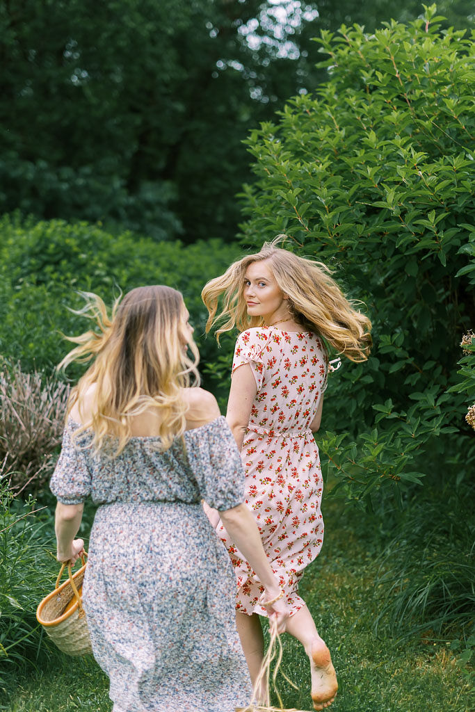 Two women with long blonde hair, wearing red and blue summer dresses, run barefoot through a forest. Photographed for Verde Lusso by Washington DC brand photographer Kim Branagan.