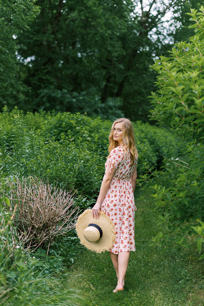 A woman wearing a white and red summer sundress holds a wide-brimmed straw hat in her hand. Her back is to the camera, but she is has turned her head to look back at the camera. She is walking through a forest.