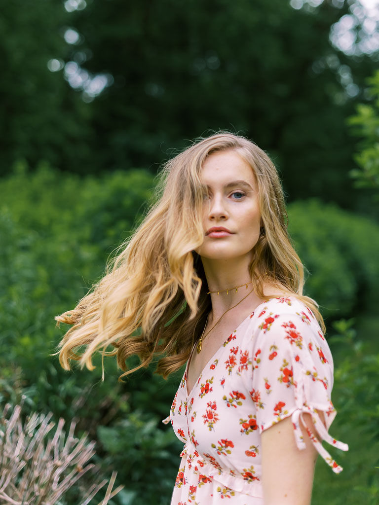 A woman stares at the camera mid-turning her body, so her hair is flowing in the air. She is blonde, has blue eyes, and is wearing a white sundress with red flowers on it. She is standing in a park with lots of green trees behind her. Photographed for Verde Lusso by Washington DC brand photographer Kim Branagan.