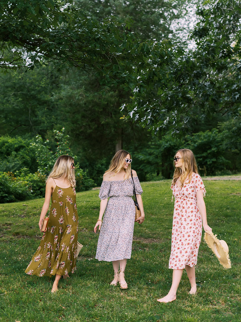 Three women wearing colorful, flowy sundresses stand and chat with each other in a park. A woman stares at the camera mid-turning her body, so her hair is flowing in the air. She is blonde, has blue eyes, and is wearing a white sundress with red flowers on it. She is standing in a park with lots of green trees behind her. Photographed for Verde Lusso by Washington DC brand photographer Kim Branagan.