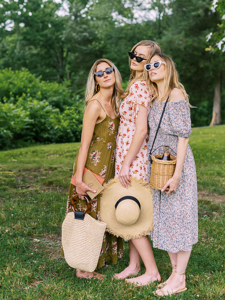 Close up shot of three women, who are wearing flowy sundresses and sunglasses, stand barefoot in a park, posing close together. They are holding straw hats and wicker tote bags. Photographed for Verde Lusso by Washington DC brand photographer Kim Branagan.