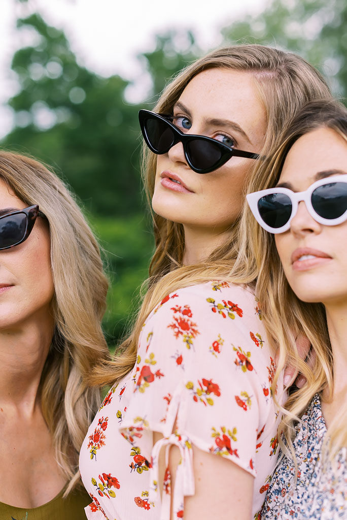 Three blonde women stare at the camera, posing close together. They are wearing colorful summer dresses, standing in a park. Photographed for Verde Lusso by Washington DC brand photographer Kim Branagan.
