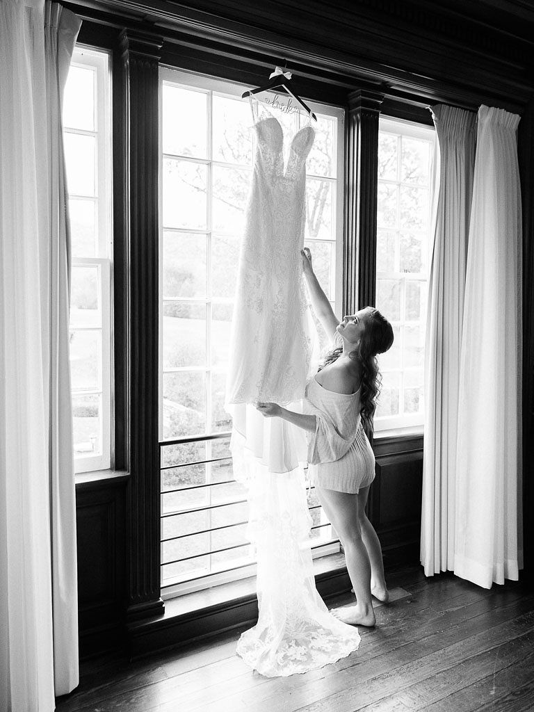 A bride touches her wedding dress, which is hanging over a window in a bedroom