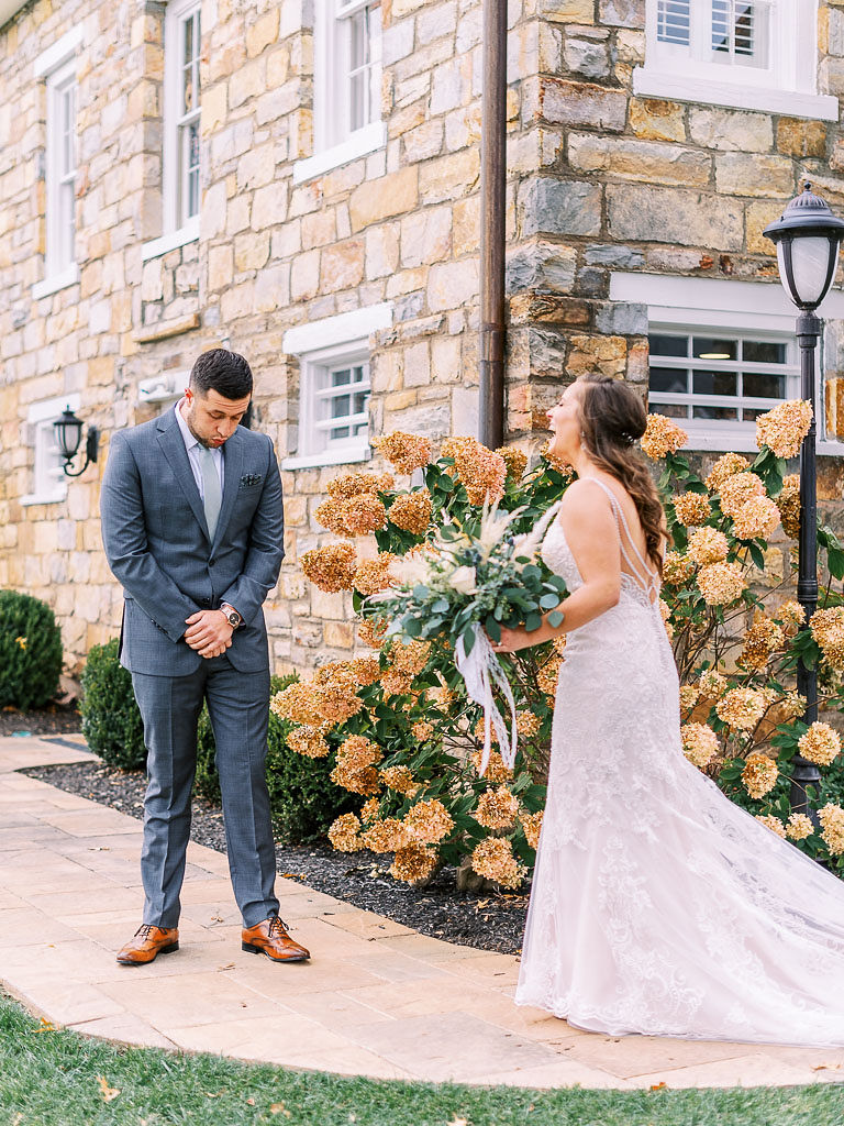 A bride walking toward her groom on their wedding day for their first look before the ceremony. The bride is smiling, and the groom has his eyes closed. Photographed by Virginia wedding photographer Kim Branagan.