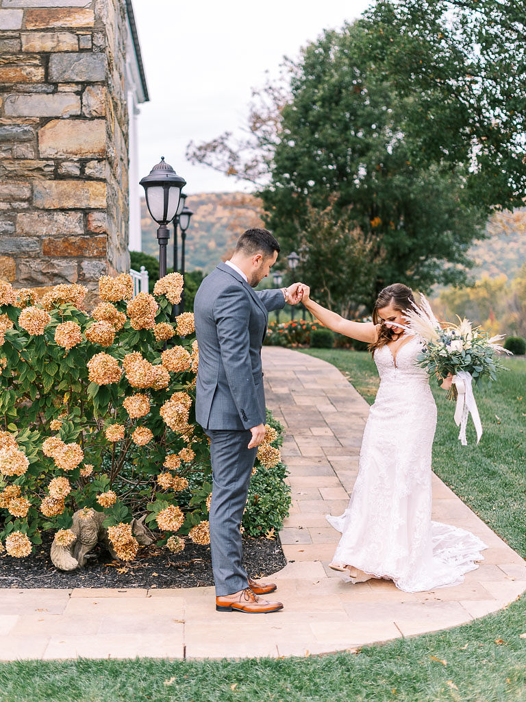 A man spins his bride around during their first look on their wedding day. They are standing next to a bush of lush, blooming flowers.