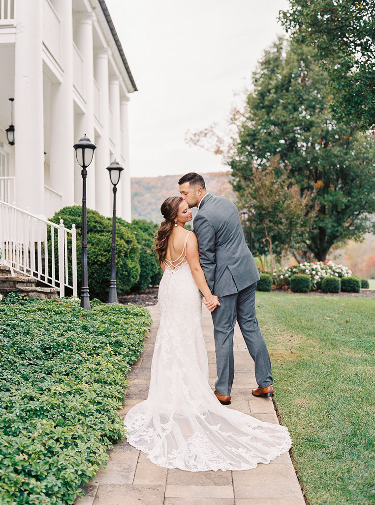 A man and woman walk on a brick path with their backs to the camera. It's their wedding day. The man is kissing his bride on the cheek.