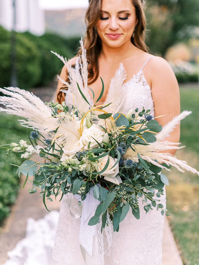 A bride hols her bridal bouquet on her wedding day. Her bouquet is comprised of lots of greenery, fall flowers, and stems of wheat. Photographed by northern Virginia wedding photographer Kim Branagan.