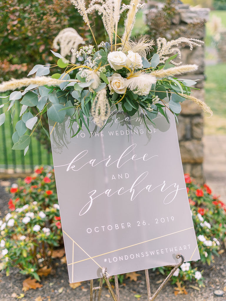 A welcome sign welcoming guests to the wedding with a lush floral arrangement on top of the sign