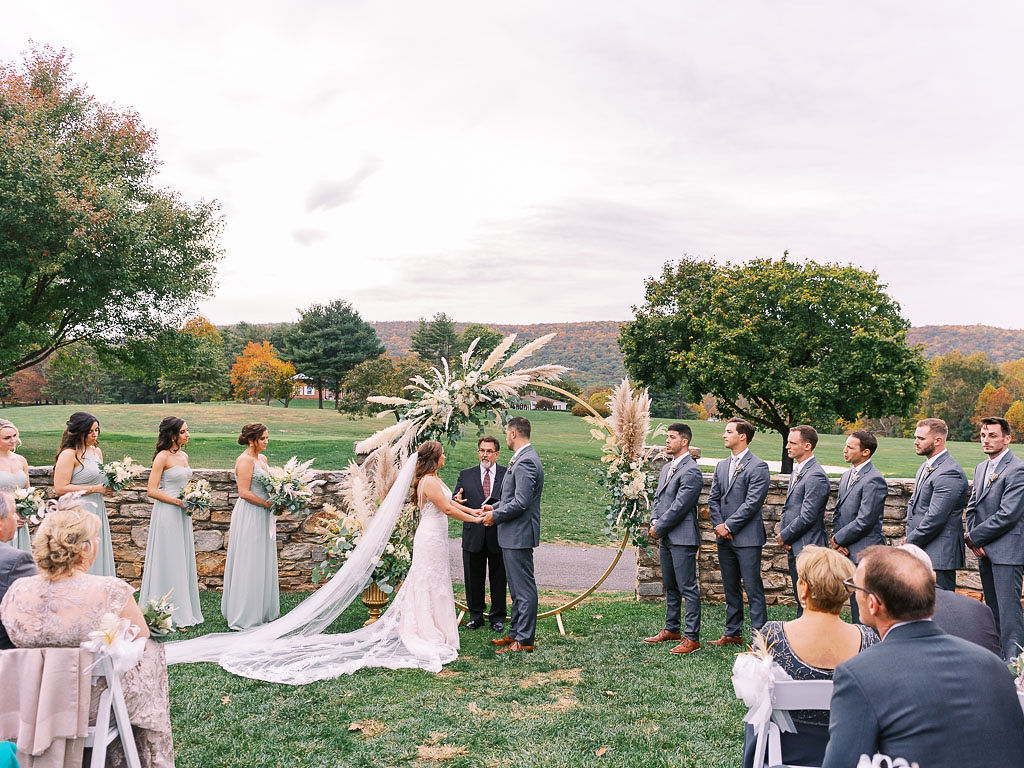 A bride and groom face each, holding hands and laughing during their wedding ceremony. They are surrounded by their wedding party. Photographed by northern Virginia wedding photographer Kim Branagan.