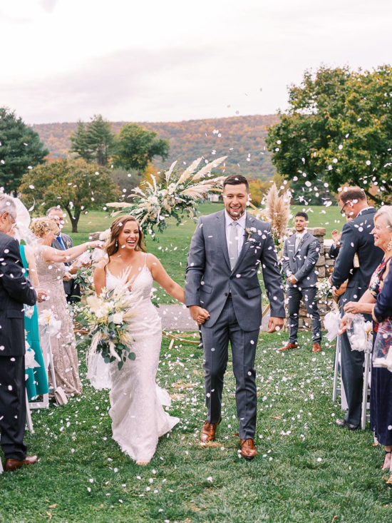 A bride and groom walk down the aisle leaving their outdoor wedding ceremony. Their friends and family are throwing confetti on them. Photographed by northern Virginia wedding photographer Kim Branagan.
