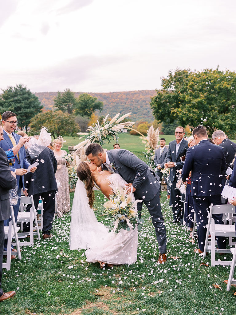 A bride and groom kiss walking down the aisle leaving their wedding ceremony. Their wedding guests are throwing confetti on them.