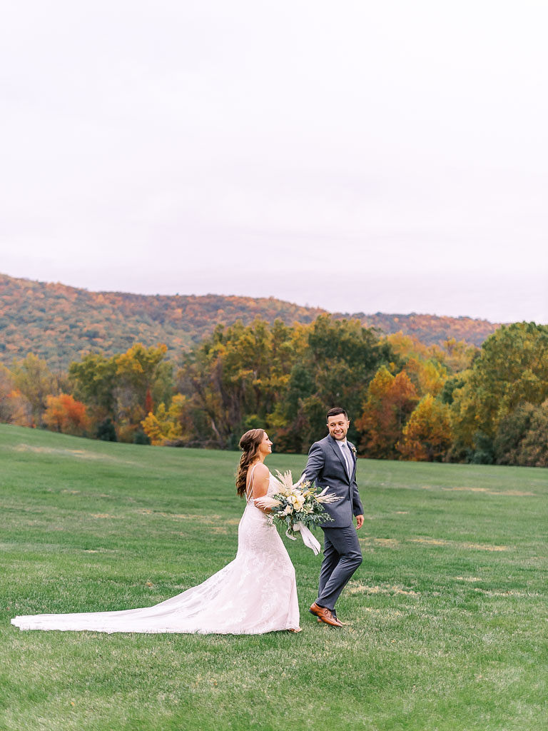A couple walks through the northern Virginia countryside, with gorgeous fall trees in the backdrop. They are holding hands, and the bride is holding her bouquet. Photographed by northern Virginia wedding photographer Kim Branagan.