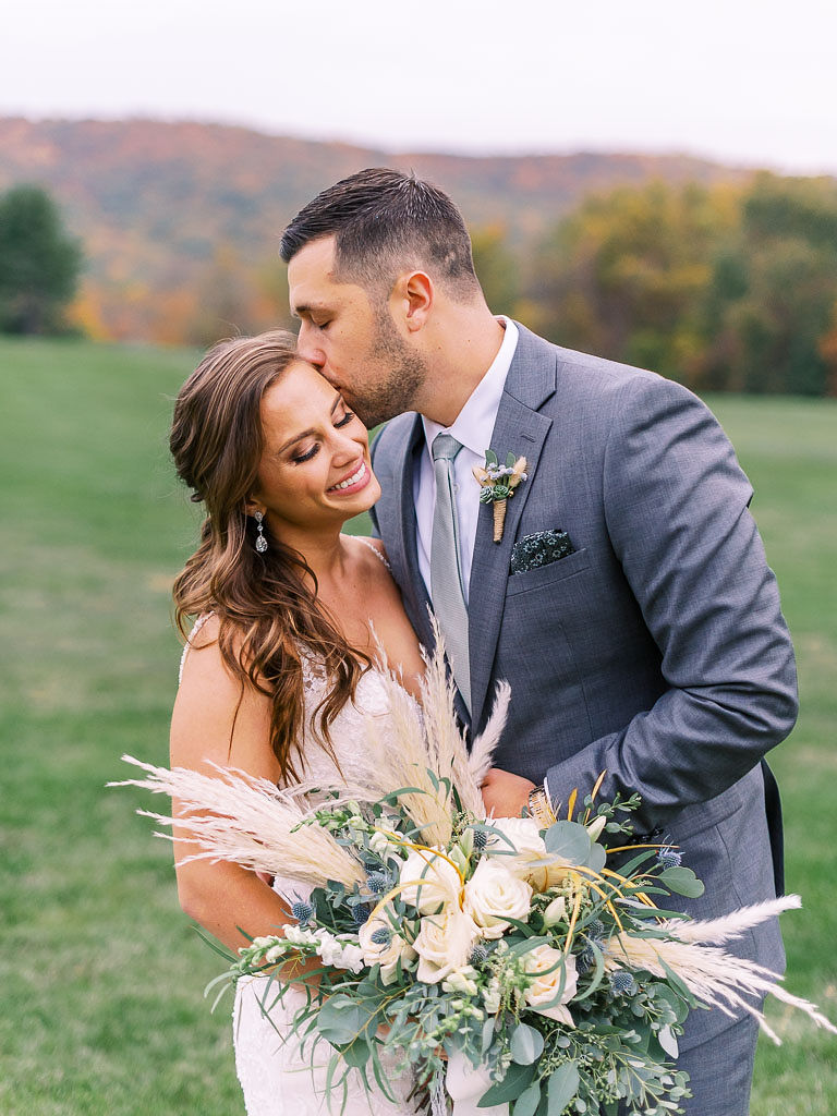 A bride smiles and closes her eyes while the groom kisses her forehead. The bride is holding her lush bridal bouquet. Photographed by northern Virginia wedding photographer Kim Branagan.